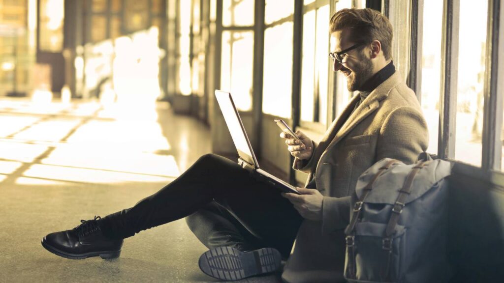 A businessman using his laptop and smartphone while sitting on the floor of the airport 